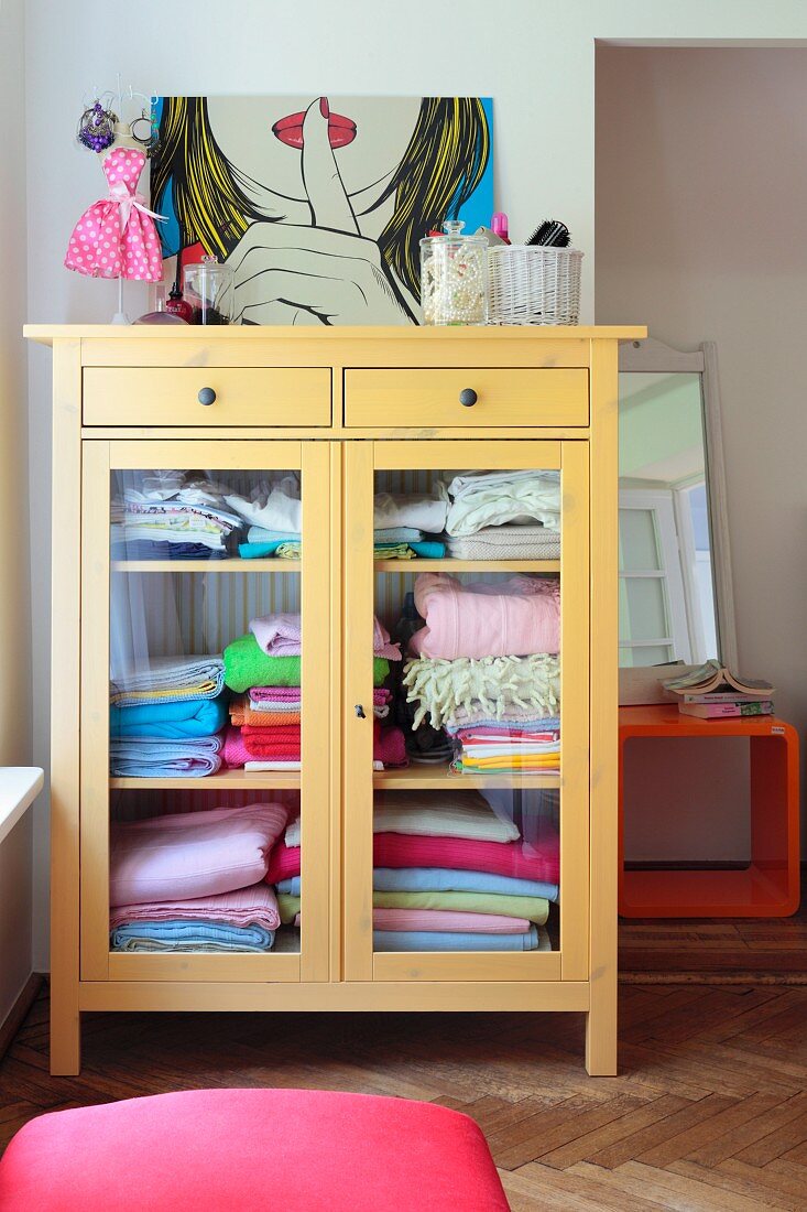 Pop-art picture leaning against wall on top of yellow-painted, half-height cabinet with glass doors