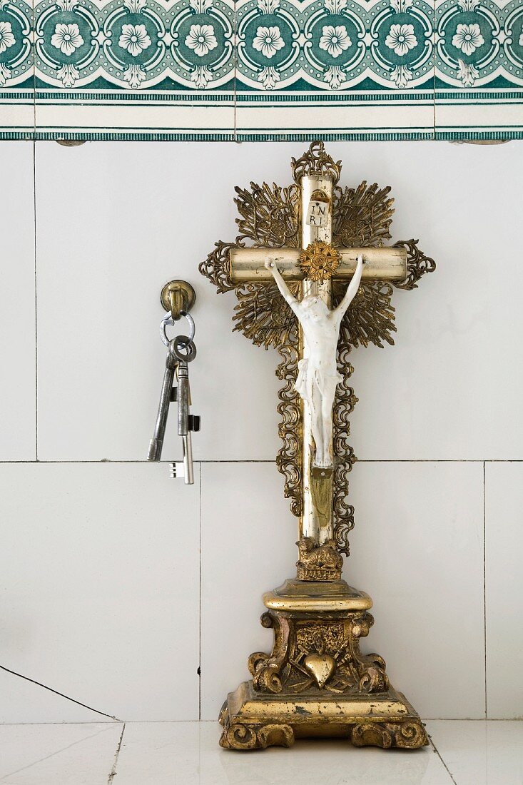 Altes, vergoldetes Stand Kruzifix und Schlüsselbund an Messinghaken vor einer alten, weiss gefliesten Wand mit Blütenbordüre