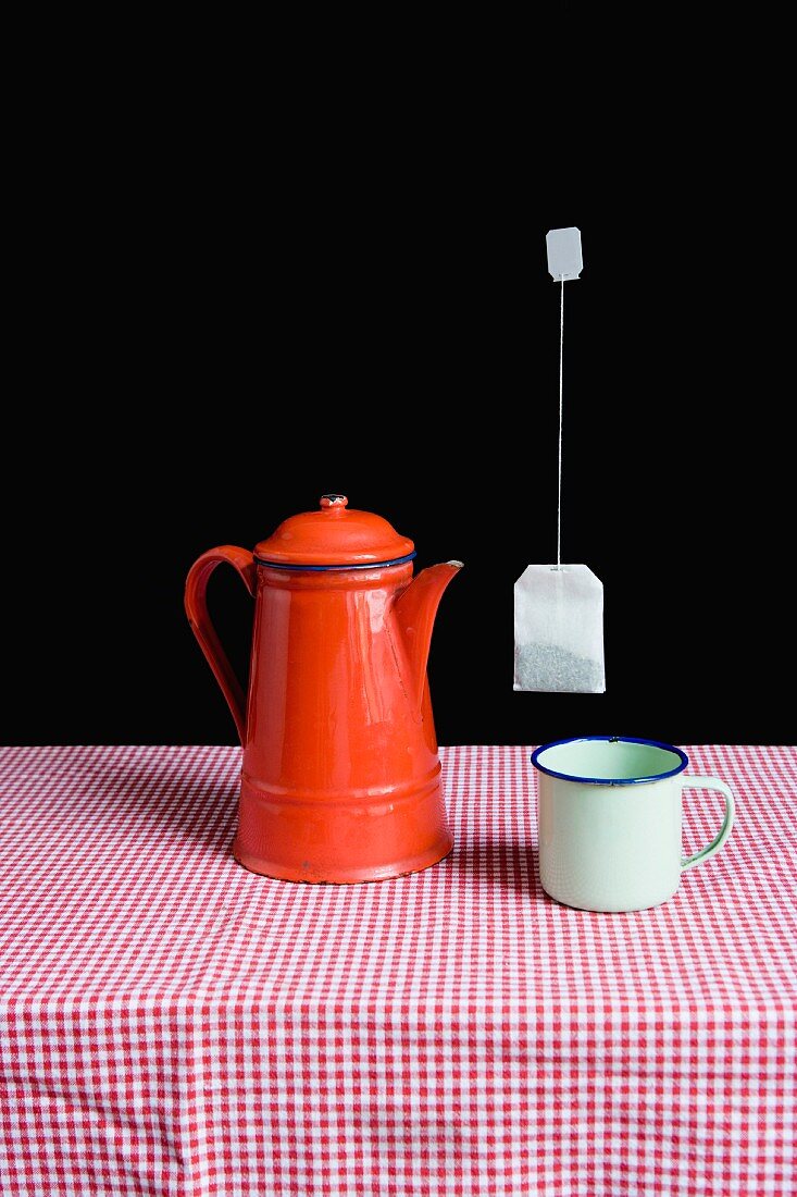 Emaille-Teekanne und Tasse auf Tisch, darüber schwebt ein Teebeutel in der Luft