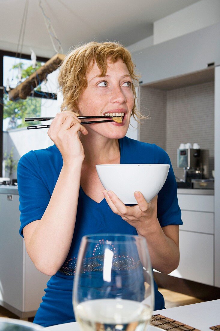 A woman eating a stir-fry with chopsticks in a kitchen