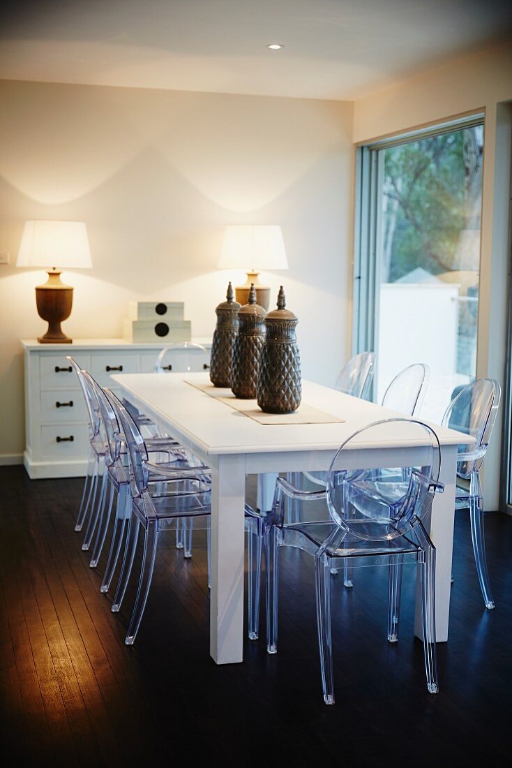 Dining room with white wooden table, plexiglass chairs, sideboard with drawers and lamps