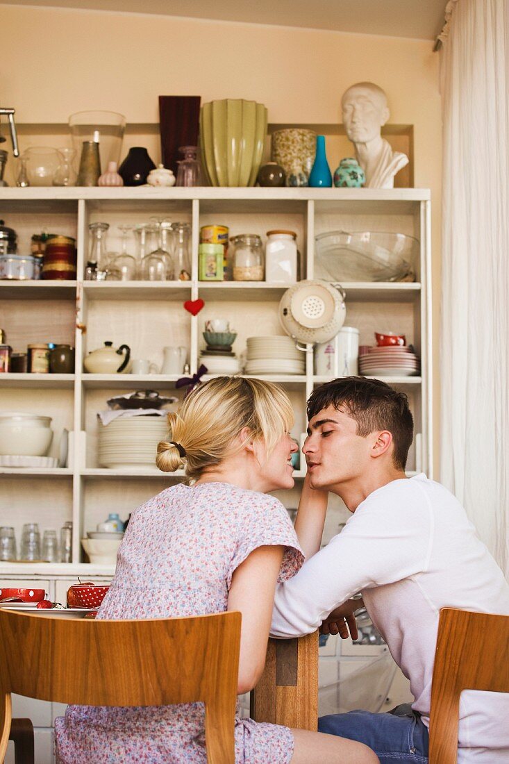 A young couple flirting at a kitchen table with a shelf of crockery in the background