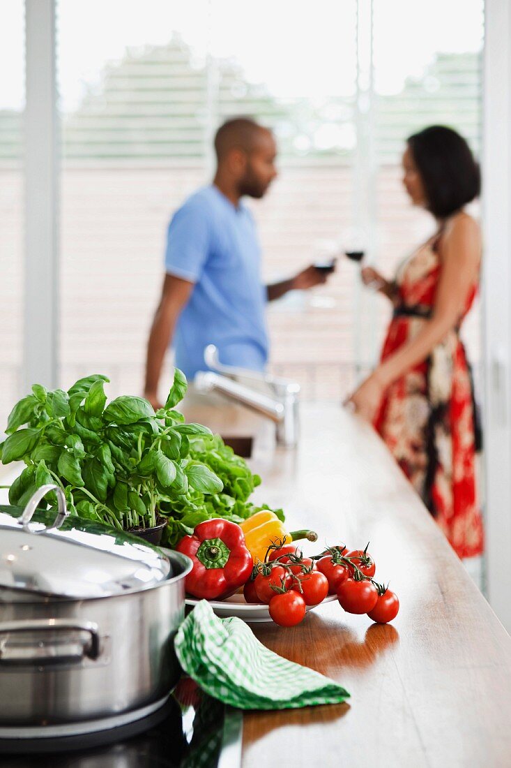 Vegetables and herbs and a pot on a work surface with a young couple in the background