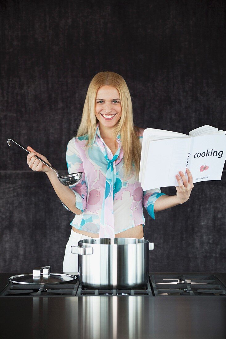 A happy woman with a cookbook and a ladle in front of a stove