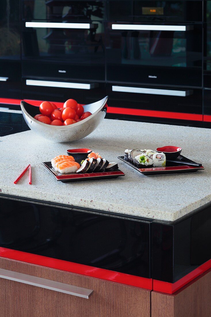 Ausschnitt eines Küchenblockes mit schwarzen und roten Farbakzenten, auf heller Steinplatte Schale mit Tomaten und Sushi Speisen