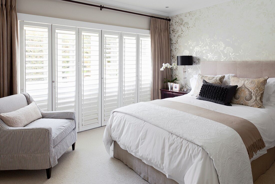 Box spring bed with headboard and scatter cushions and armchair next to terrace doors with closed interior shutters