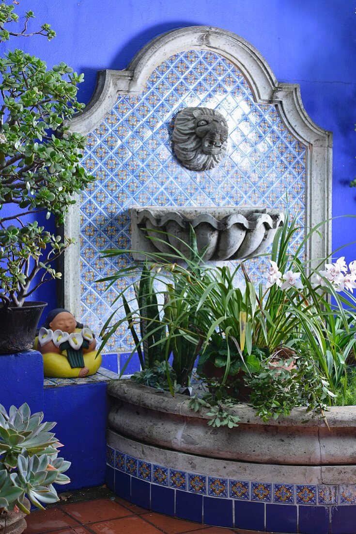 Flowering plants in semicircular raised bed with stone surround against tiled wall panel with water spout and basin in Oriental style