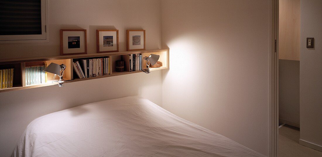 Narrow bookshelf of light wood above a bet with white linen