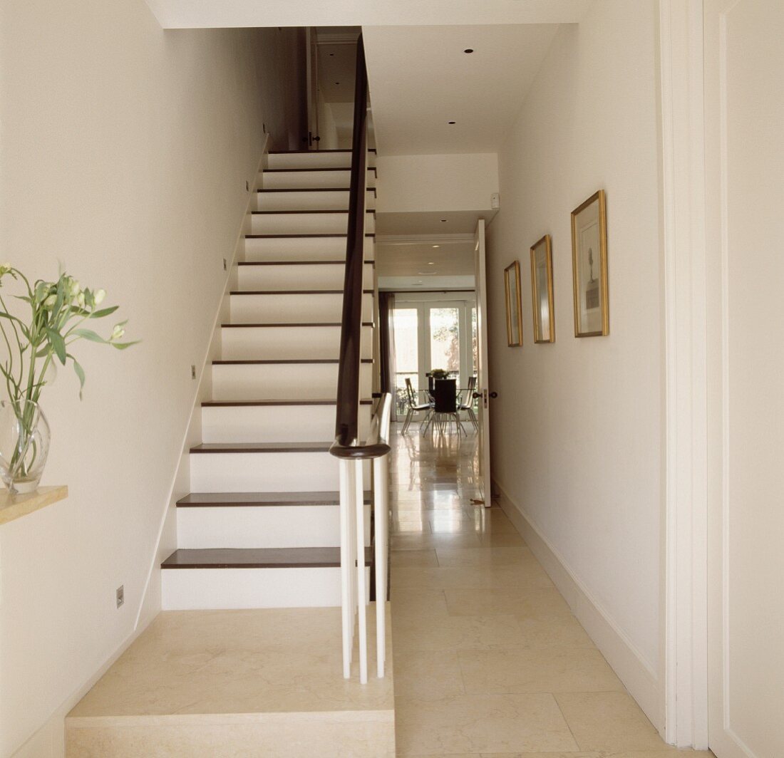 Narrow, white lacquered wooden staircase and dark railing in a stairwell with open door and view into a dining room