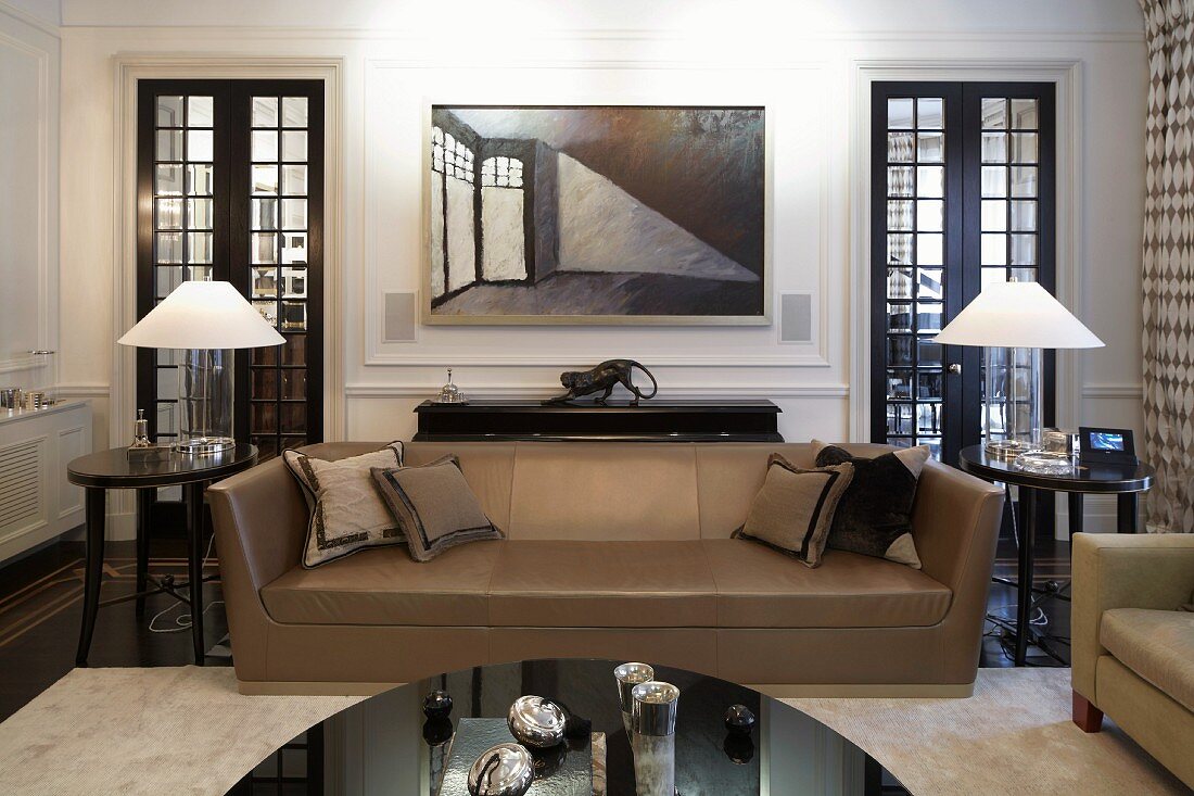 Light brown leather sofa in Art Deco style with side tables and table lamps with white shades in front of wood and glass doors