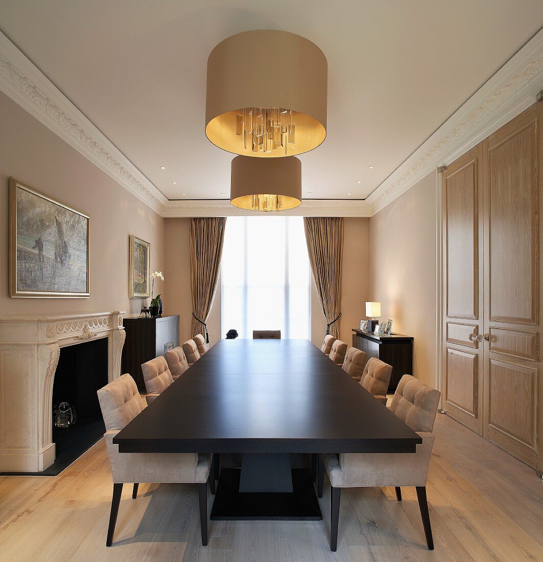 Long dining table with dark surface and upholstered chairs under a hanging lamp with gold shade