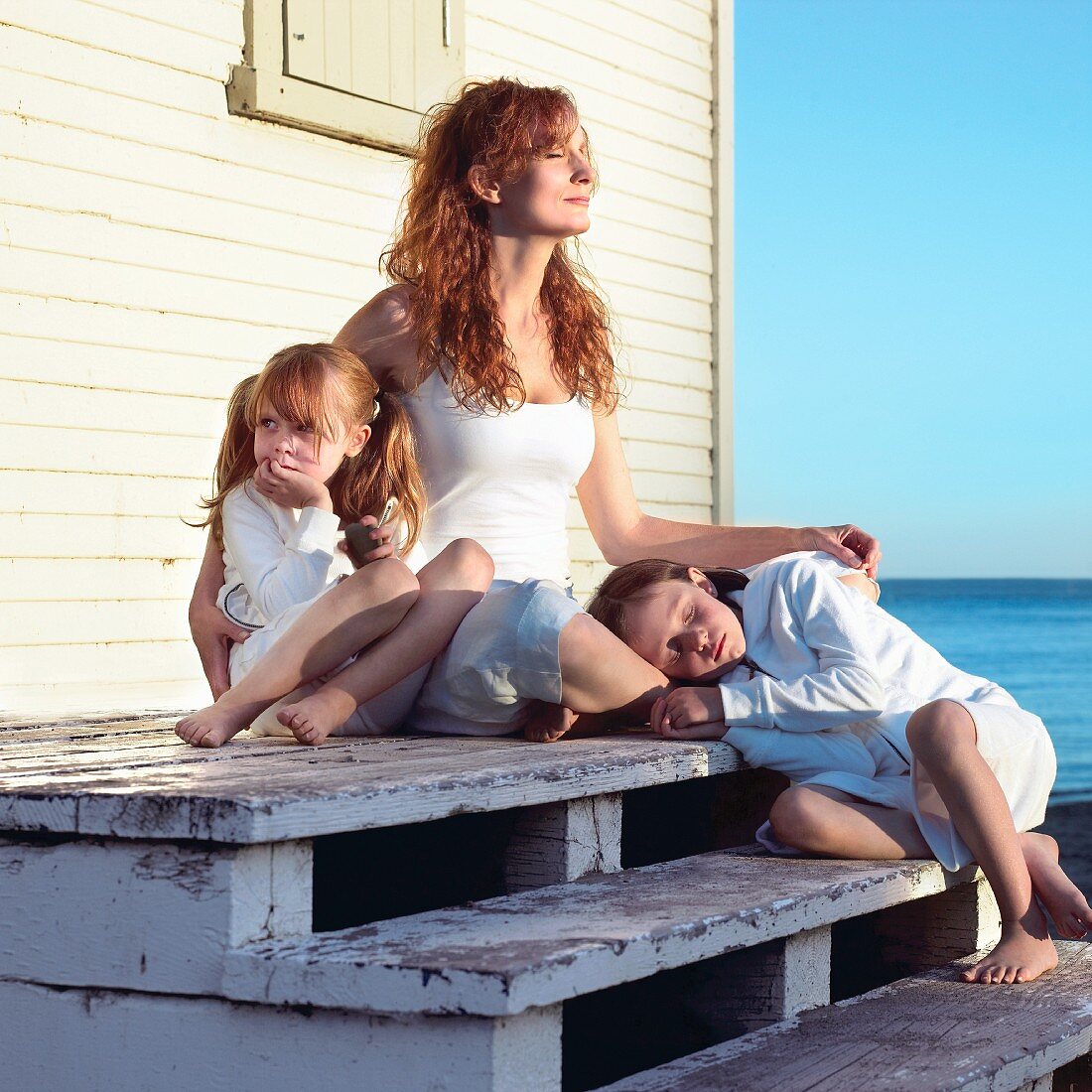 Mutter mit zwei Töchtern in der Sonne vor einem Strandhaus sitzend