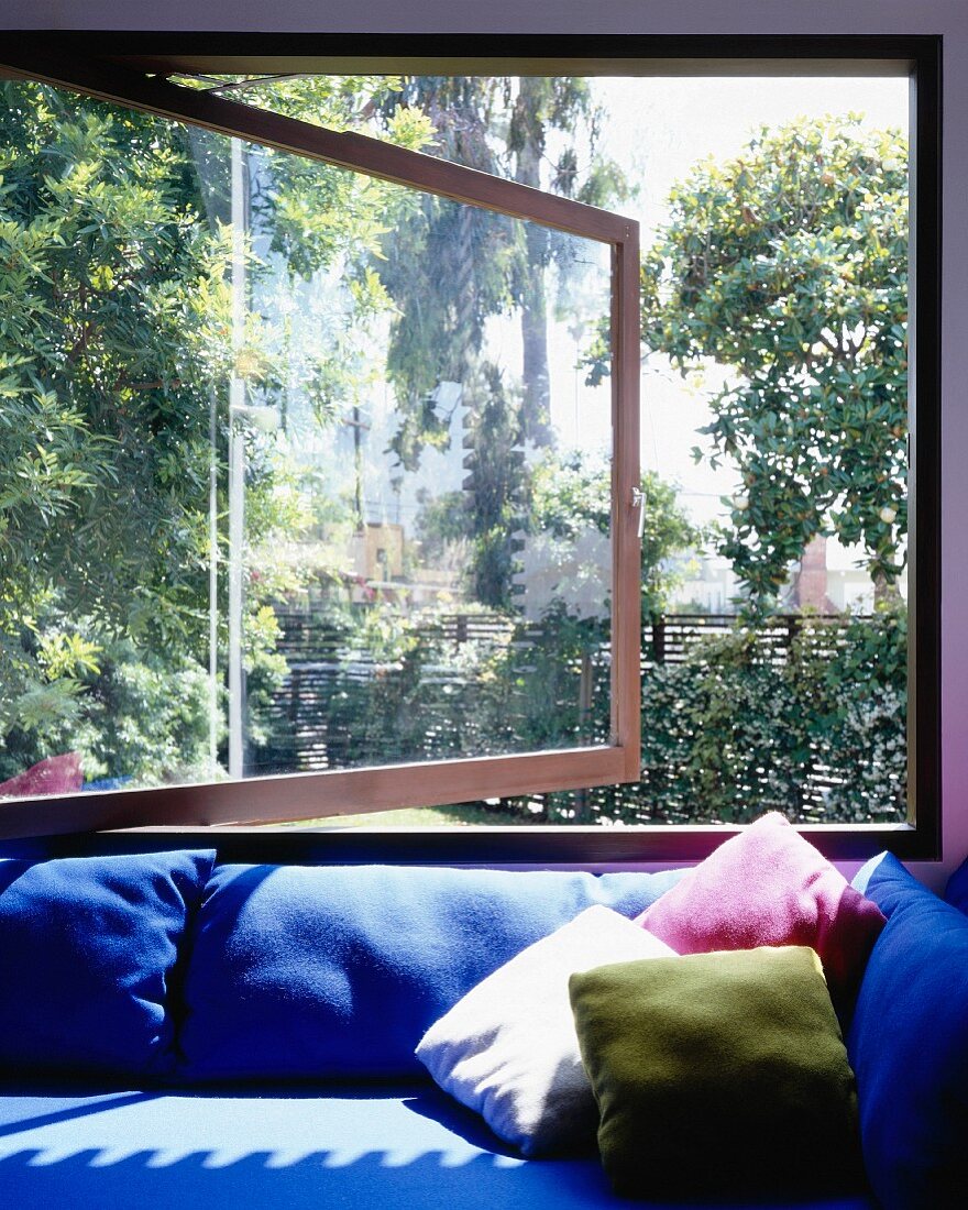 Sofa covered with blue upholstery and colorful pillow in front of an open window with a garden view
