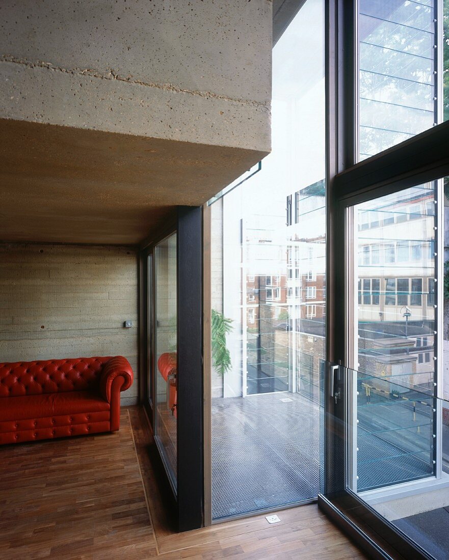 Detail of a concrete living room with brick red leather sofa and a view through the window of the balcony