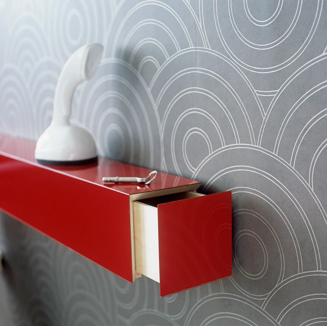 A white telephone on red shelf with an open drawer mounted on a wall hung with a geometric pattern