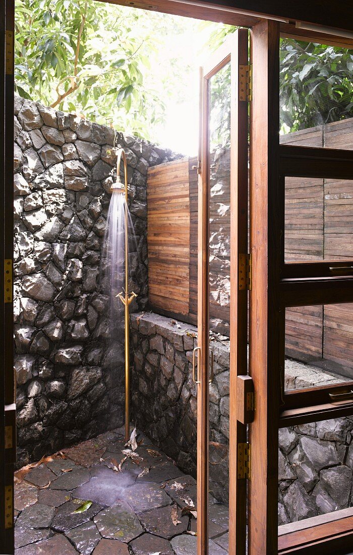 View through terrace door to vintage brass outdoor shower in corner by stone wall