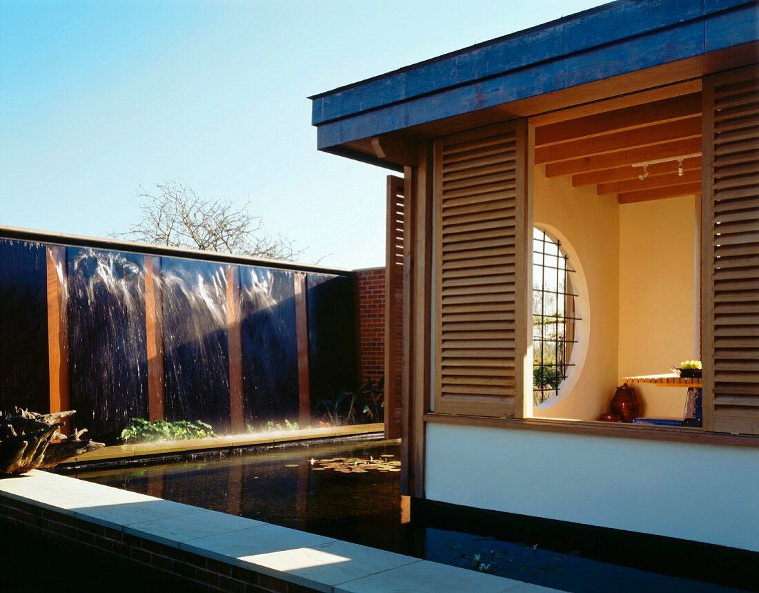 A waterfall and a pool around a modern house with a view through floor-to-ceiling windows into a living room