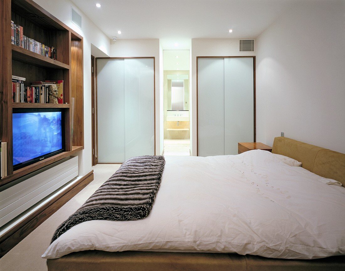 Wall cabinet with TV on opposite double bed in modern bedroom with ensuite bathroom