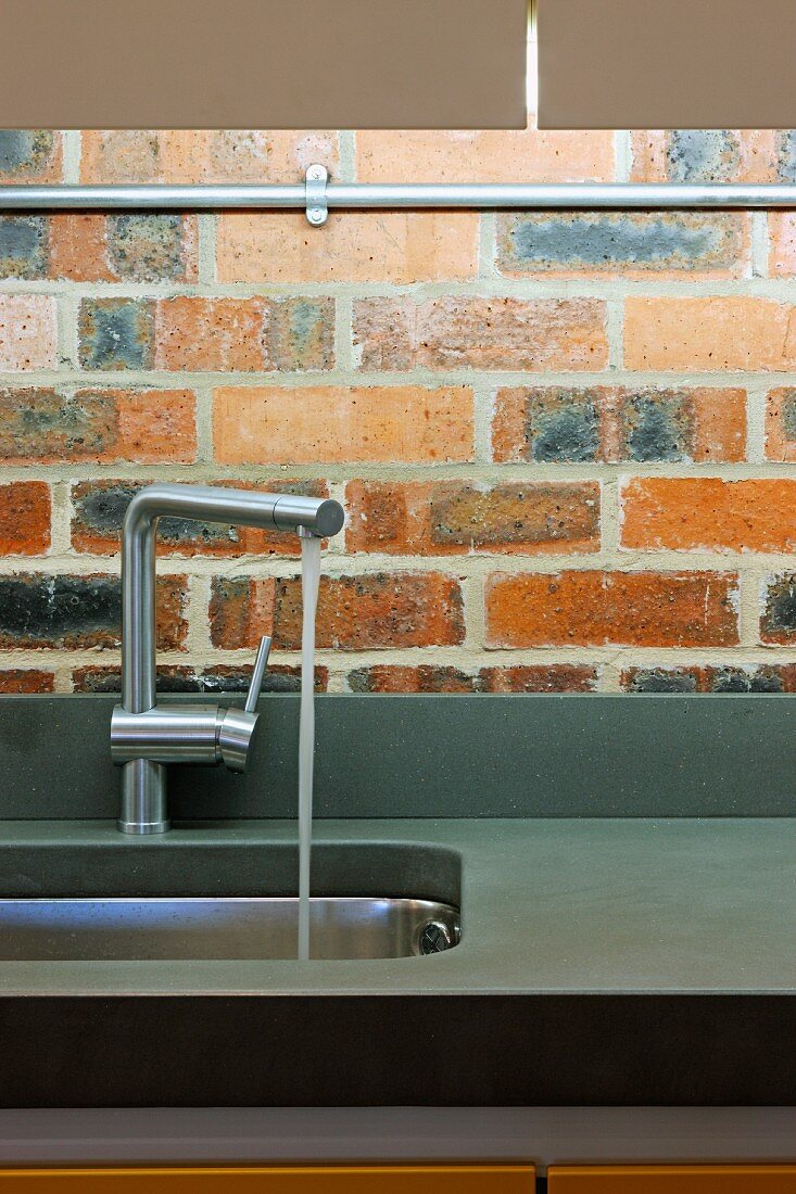 Designer Armatur mit einlaufendem Wasser in Edelstahlbecken vor Ziegelwand