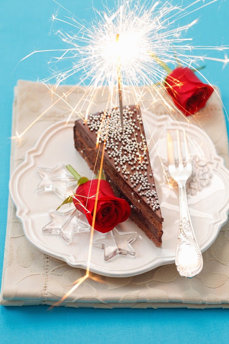 A slice of chocolate cake decorated with a sparkler and red roses