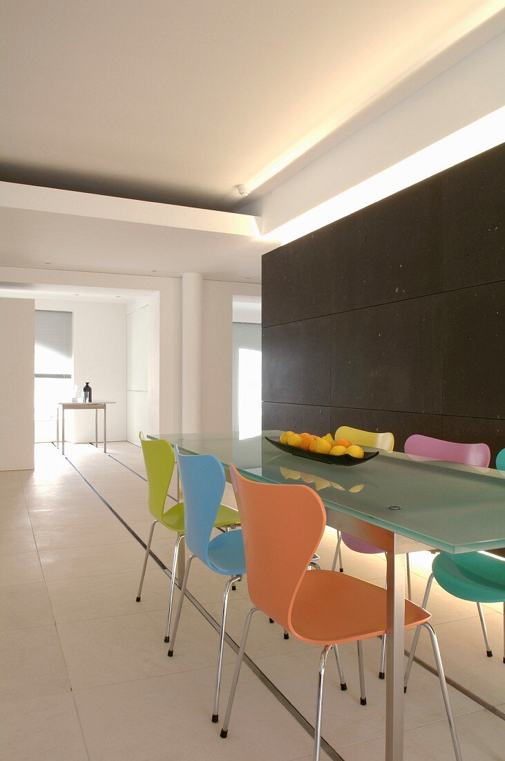 Steel and glass table and colourful 50s designer chairs in front of dark partition in purist loft; table legs on metal rails in white flooring