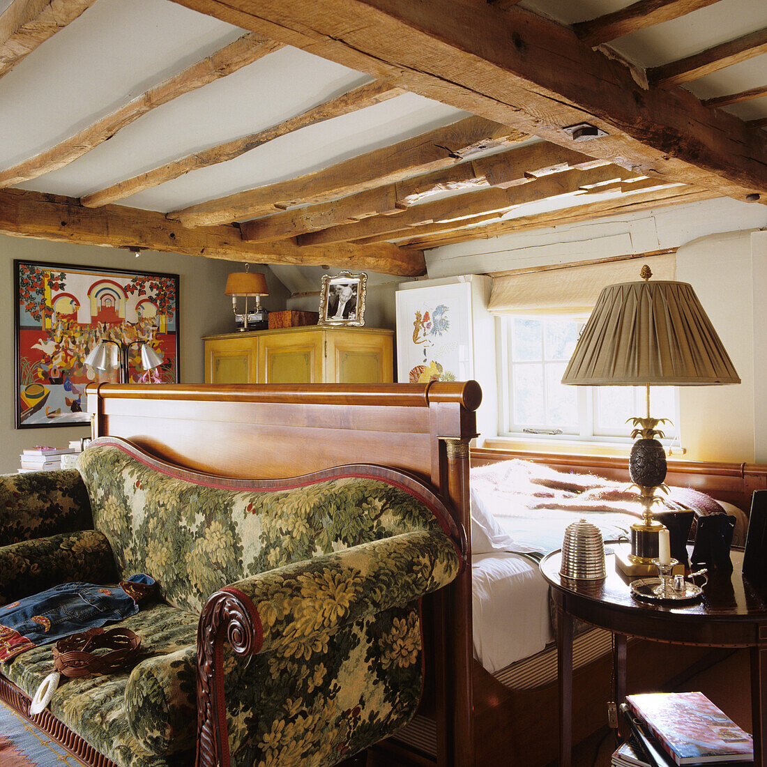 Wooden bed and vintage sofa with floral pattern in rustic bedroom with wooden beamed ceiling