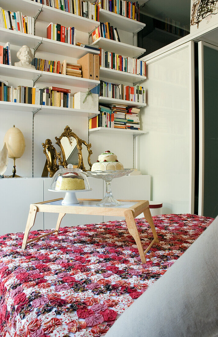 Modern bedroom with wall shelf, floral bedspread and small wooden table with biscuits