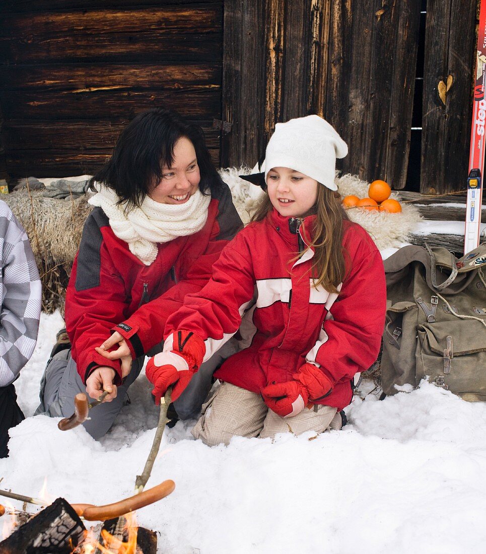 A mother and daughter grilling sausages on a open fire in front of a mountain hut