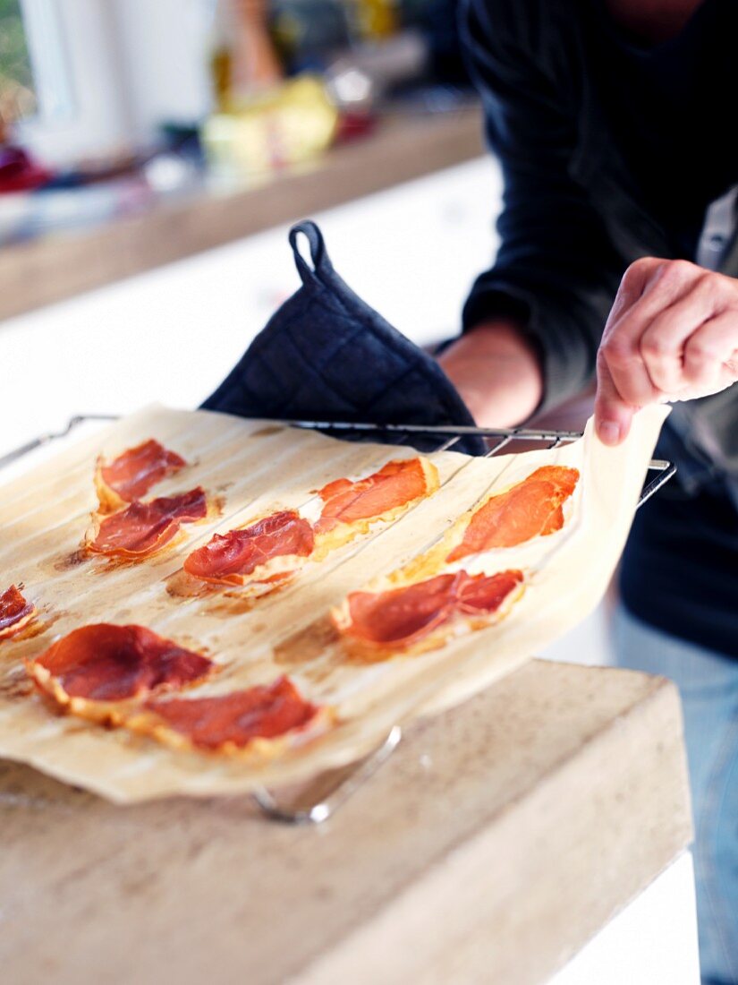 A person removing a sheet of paper with ham from an oven rack