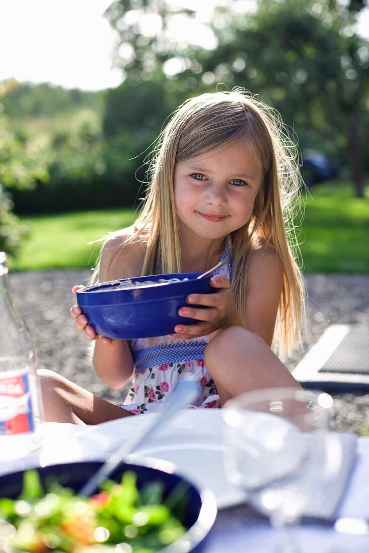 A blonde girl holding a bowl at a table laid in a garden