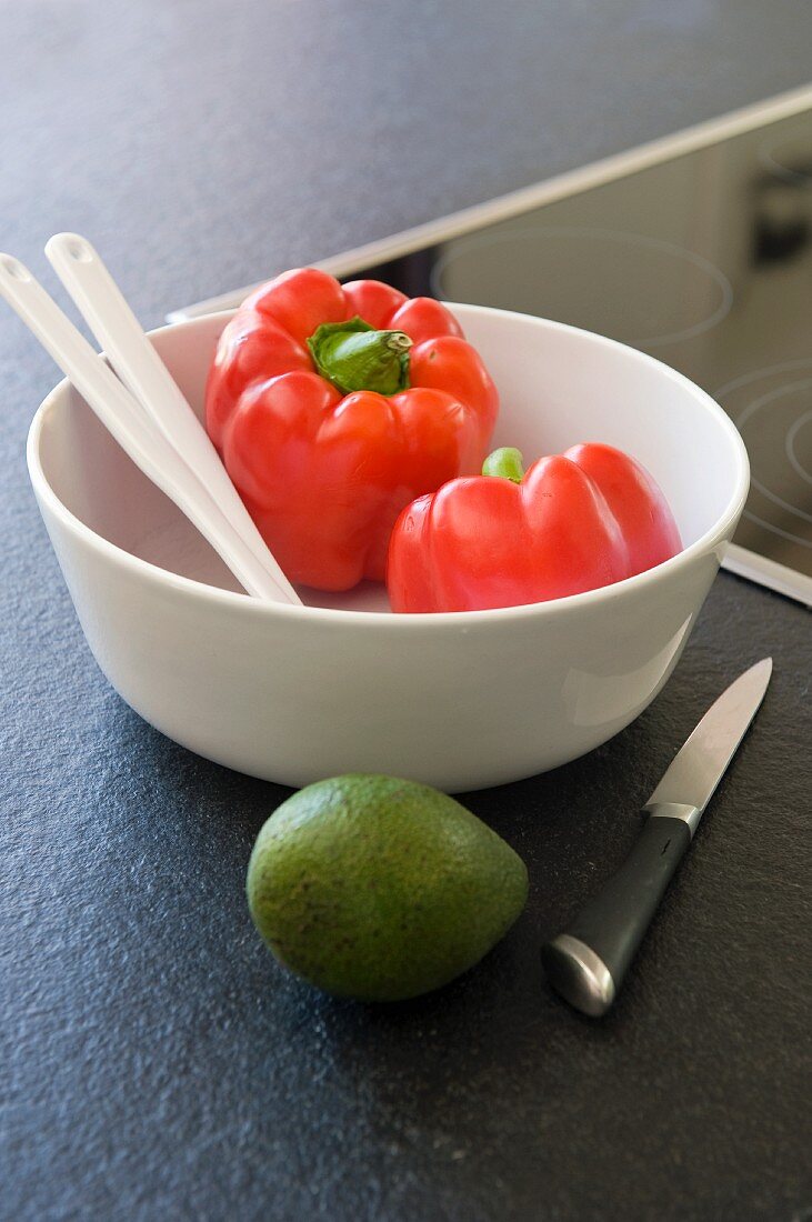 Two red peppers in bowl next to avocado