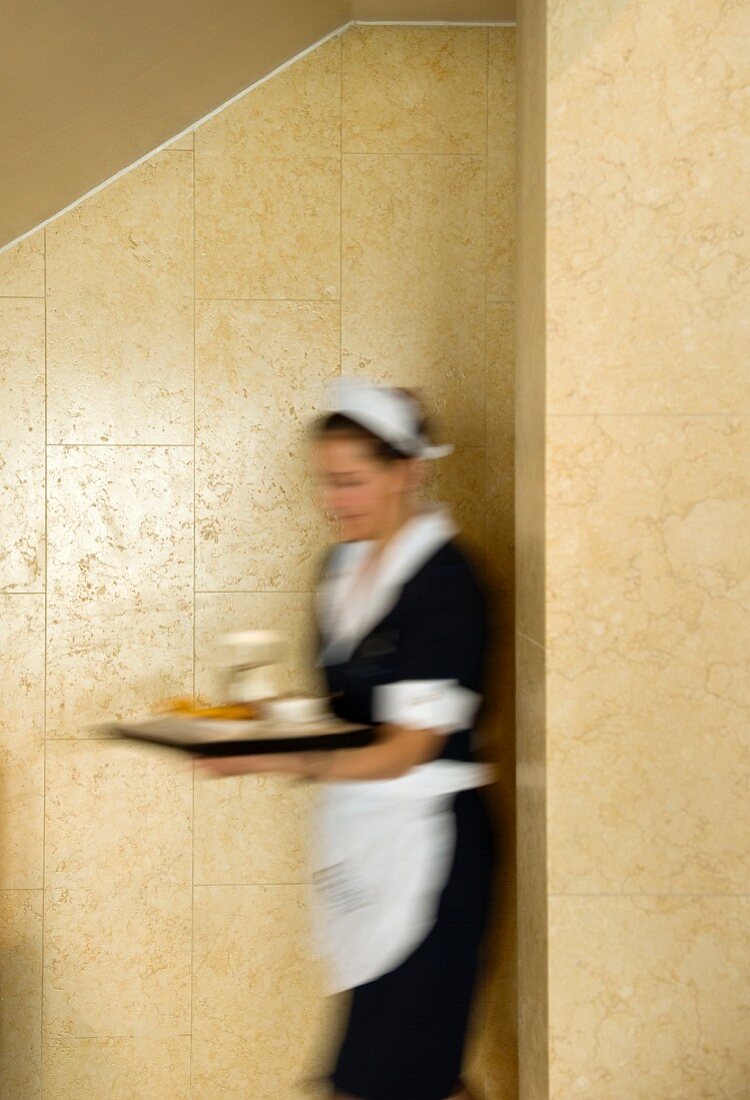 Klassisch gekleidete Kellnerin in Eile mit vollem Tablett - verschwommen vor marmorverkleideter, sandfarbener Wand