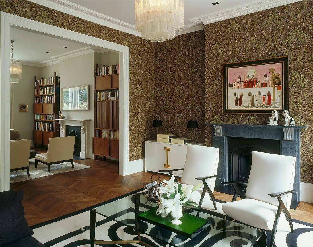 Living room with fireplace, armchairs, glass table & ornate wallpaper