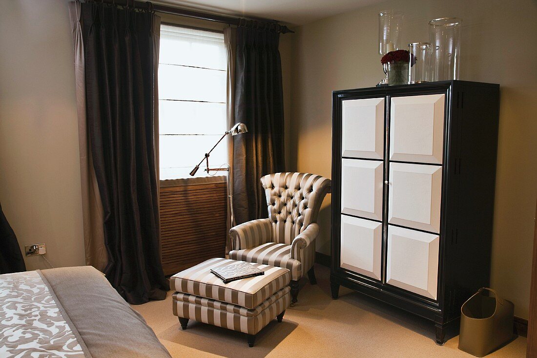 Antique armchair and footstool with matching striped covers next to black and white cupboard in bedroom