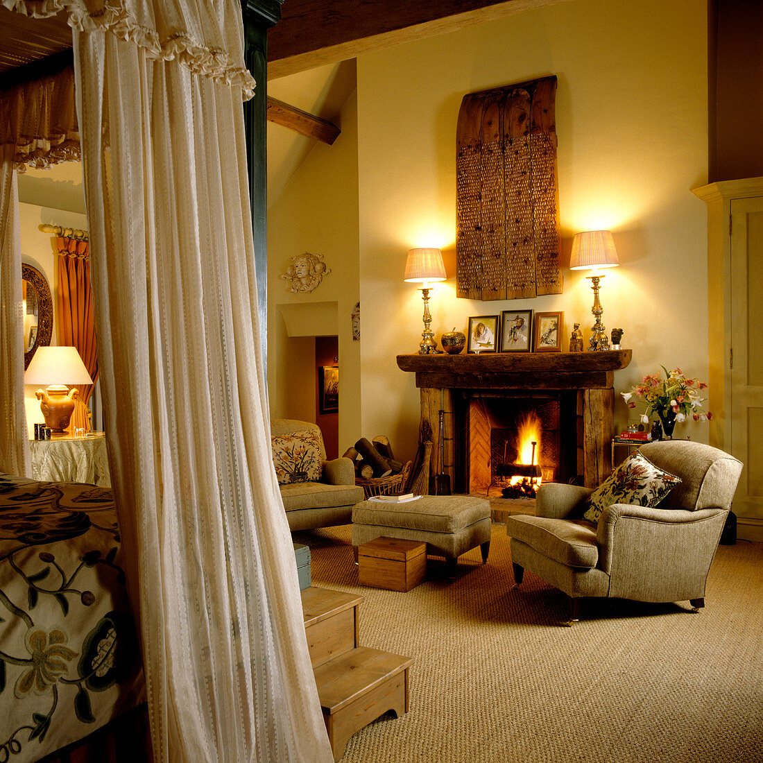 Corner of a canopied bed in front of upholstered armchairs around open fire