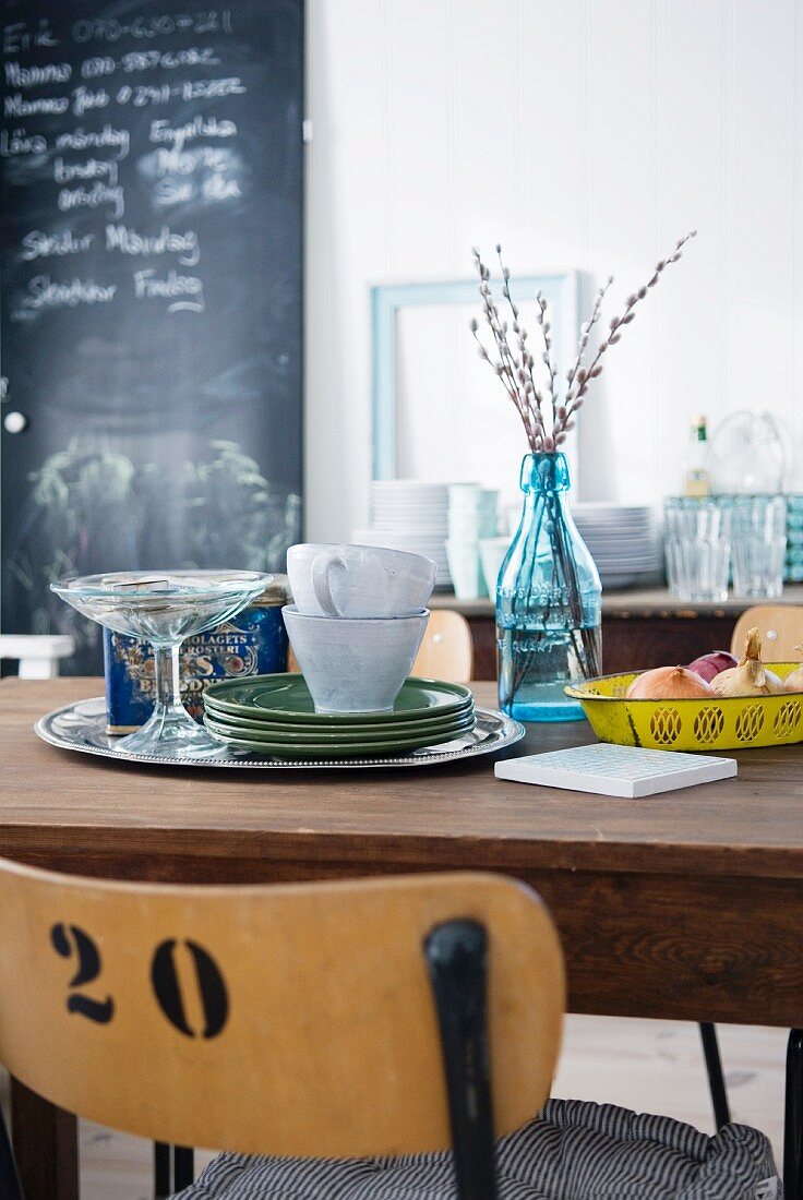 Dining room with plates and cups on wooden table, crockery on surface in background and large blackboard