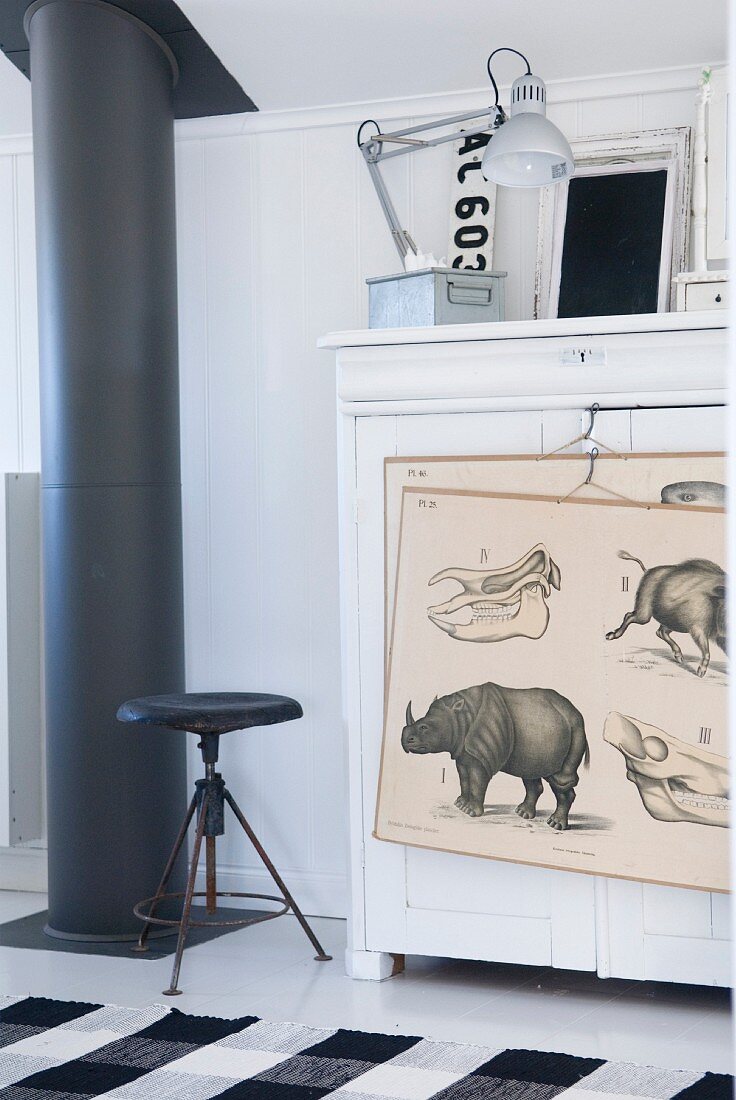 Black stove pipe and vintage stool next to white cabinet with drawings hanging on front