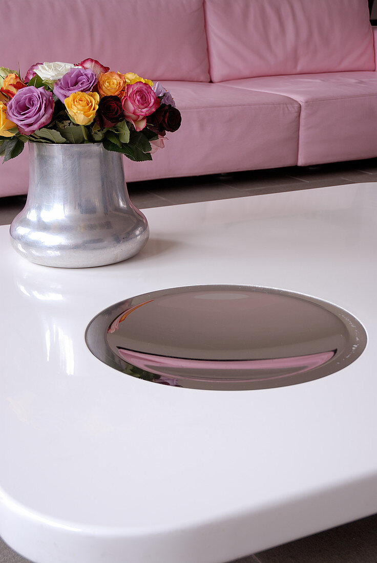 A bunch of roses and a reflective stainless steel plate on a white coffee table in front of a pink sofa