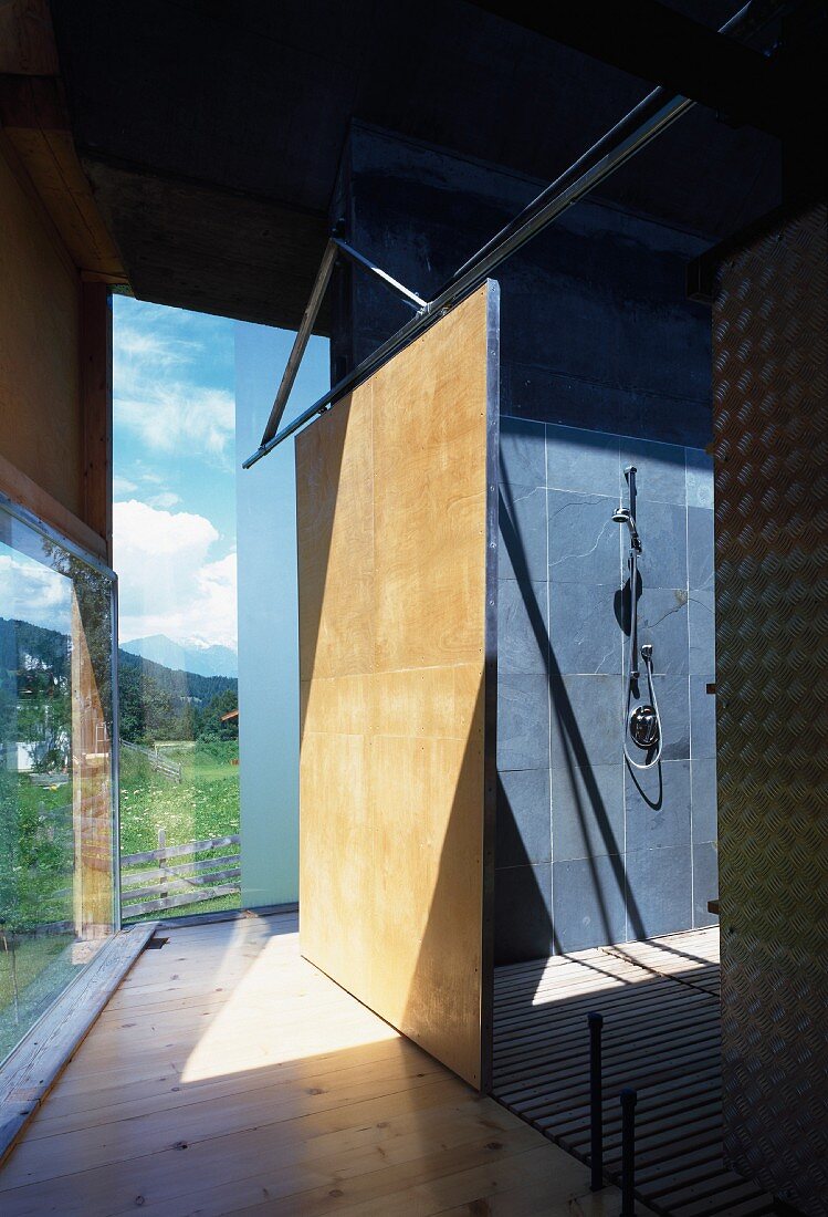 Corridor behind glass facade with view of shower room with slate grey tiling and slatted wooden floor through sliding door