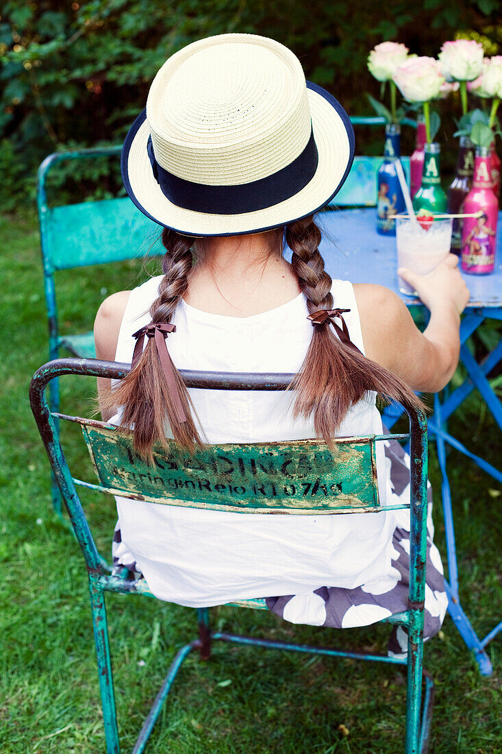 Girl with pigtails and straw hat sits on vintage garden chair in front of flower decoration