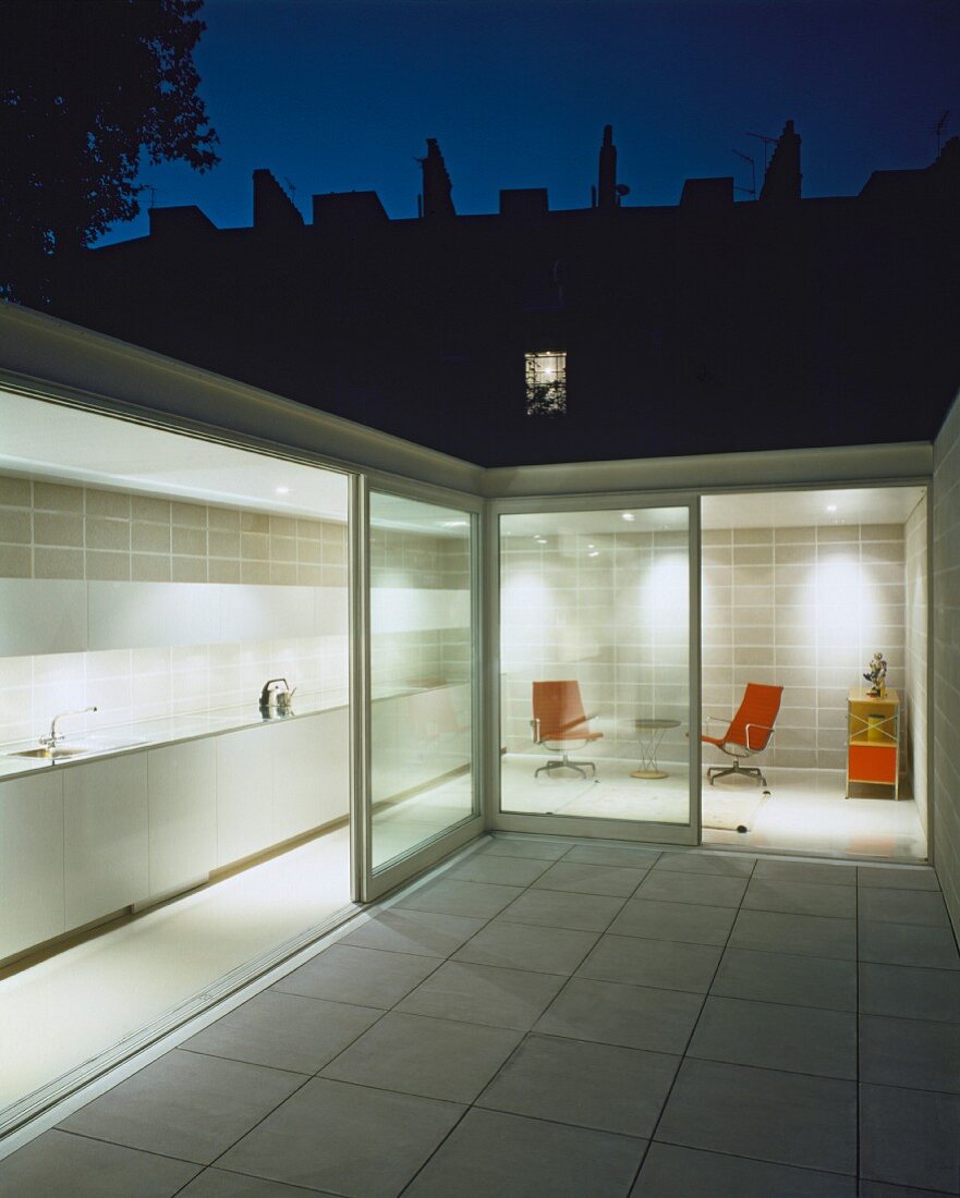 Courtyard with white tiled floor and view of illuminated kitchen and relaxation area through open sliding terrace door