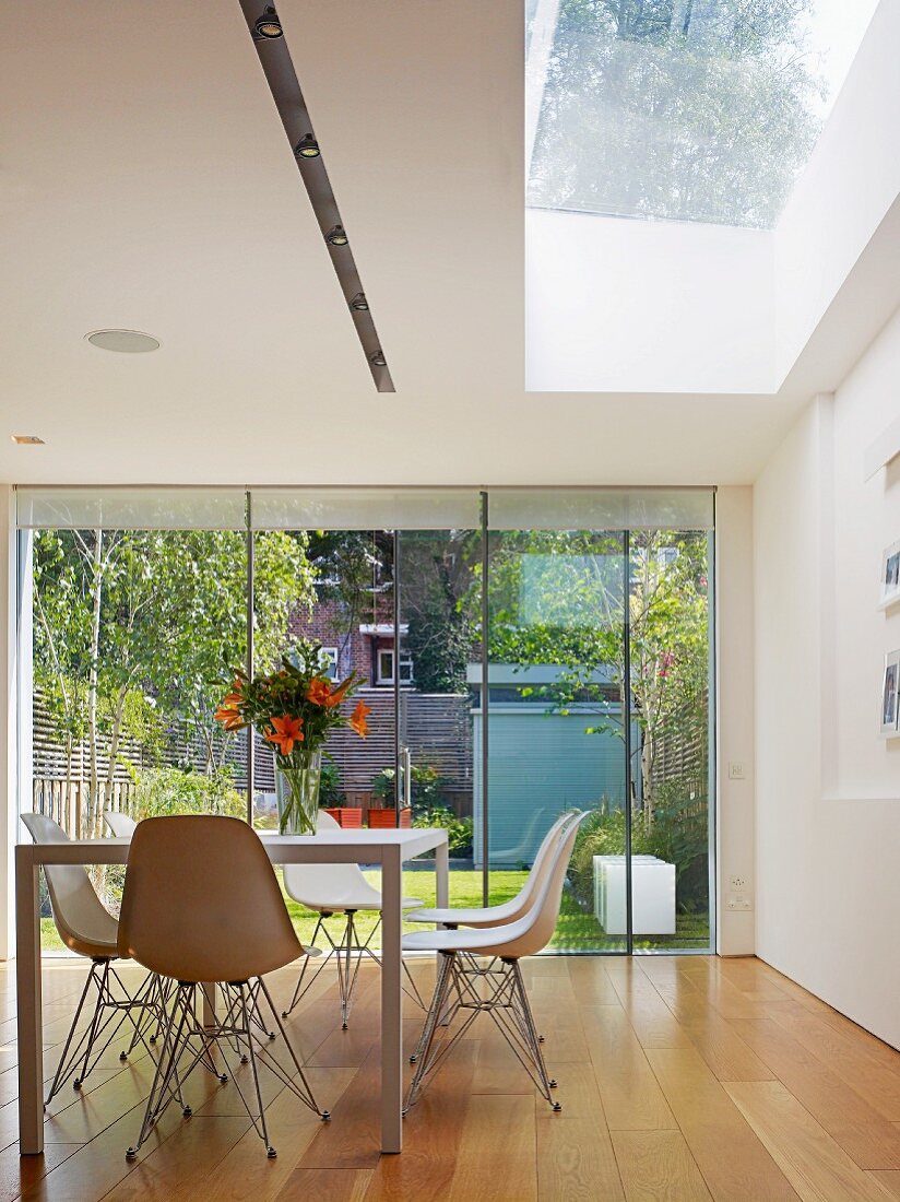 Table with white shell chairs in front of glass wall leading to terrace and beneath skylight in modern dining room