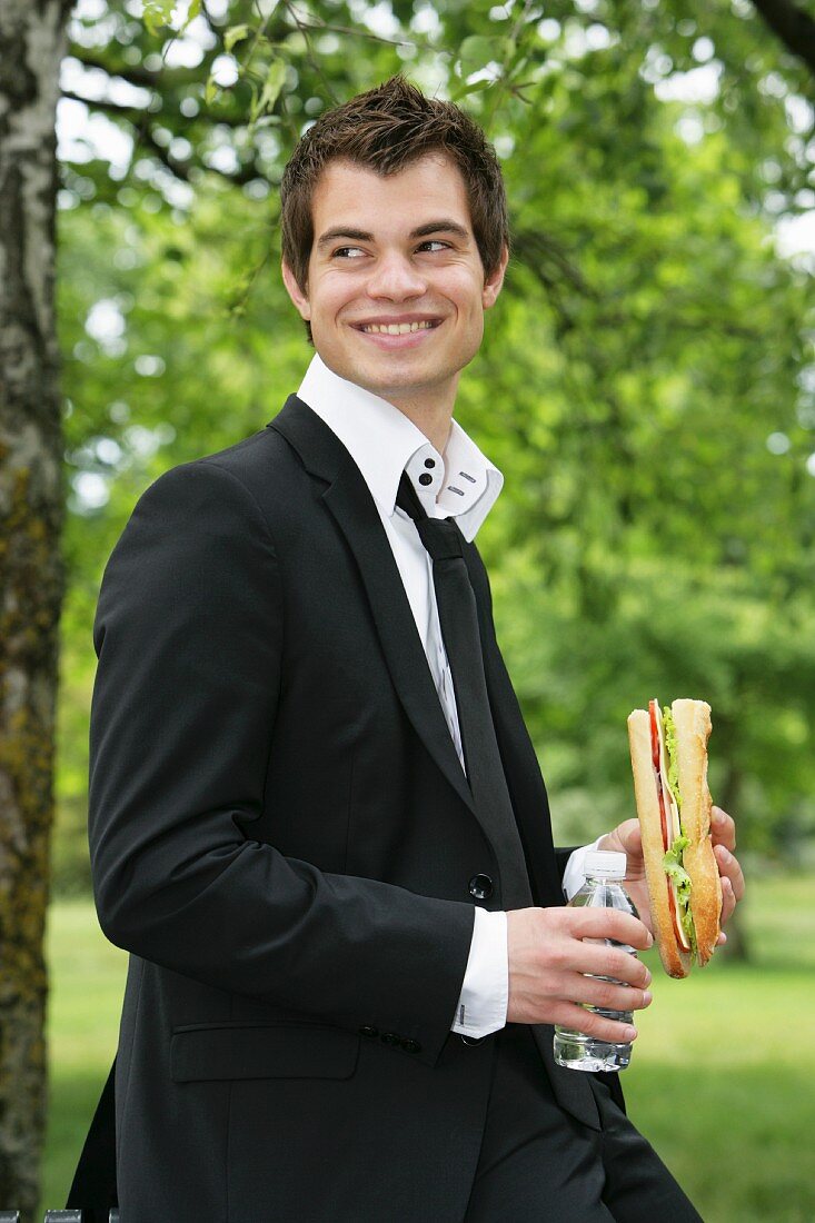 Young man with baguette sandwich and water in park