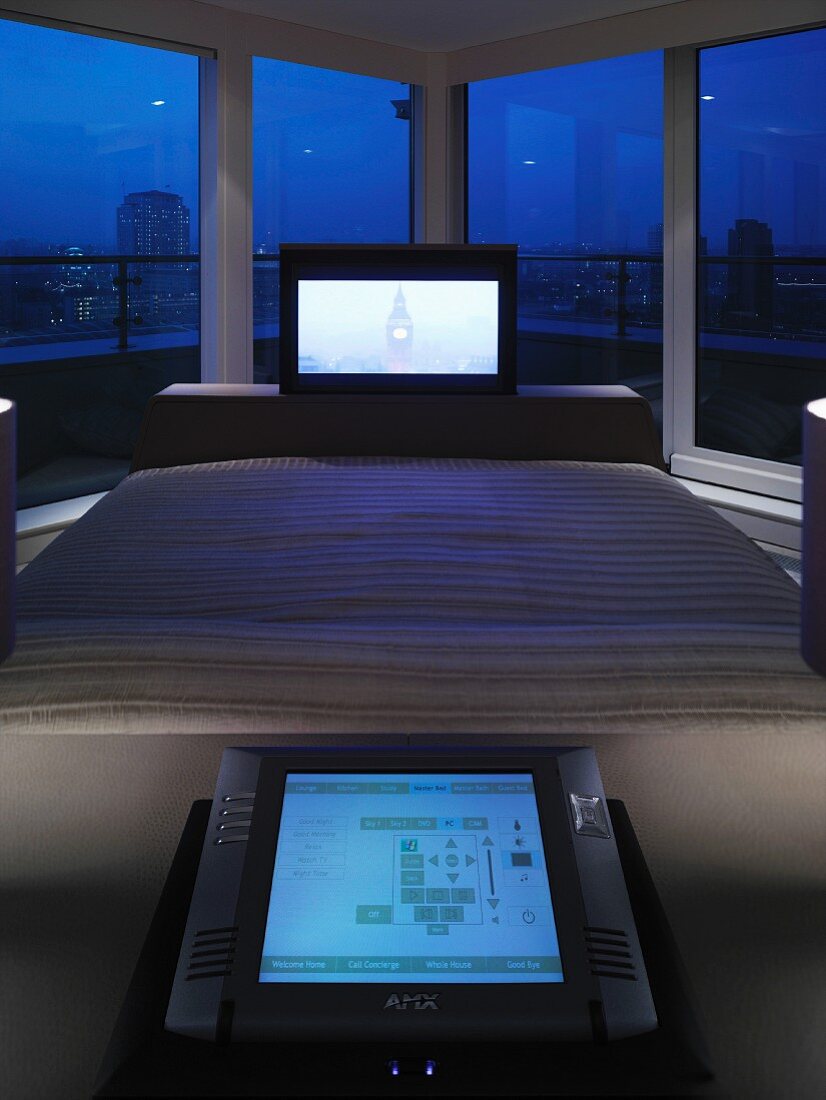 Flat-screen TV at foot of double bed in front of panoramic window with view of city at night