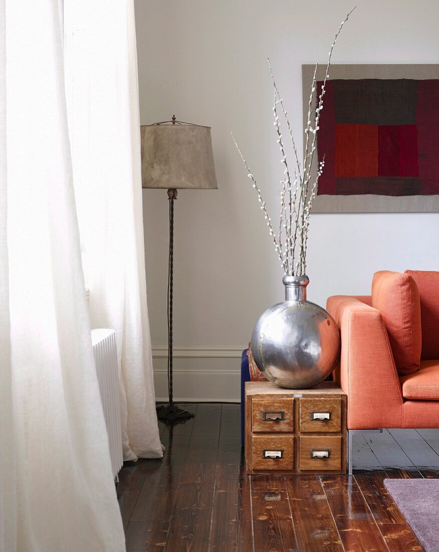 Silver-coloured vase on low, wooden cabinet and standard lamp in corner of living room