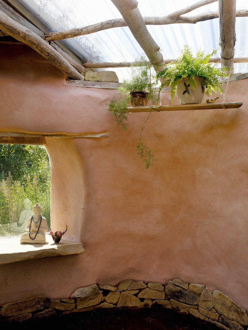 Organisch geformtes Lehmhaus mit einfacher Oberlichtkonstruktion und Buddha-Statue in der rahmenlosen Fensteröffnung