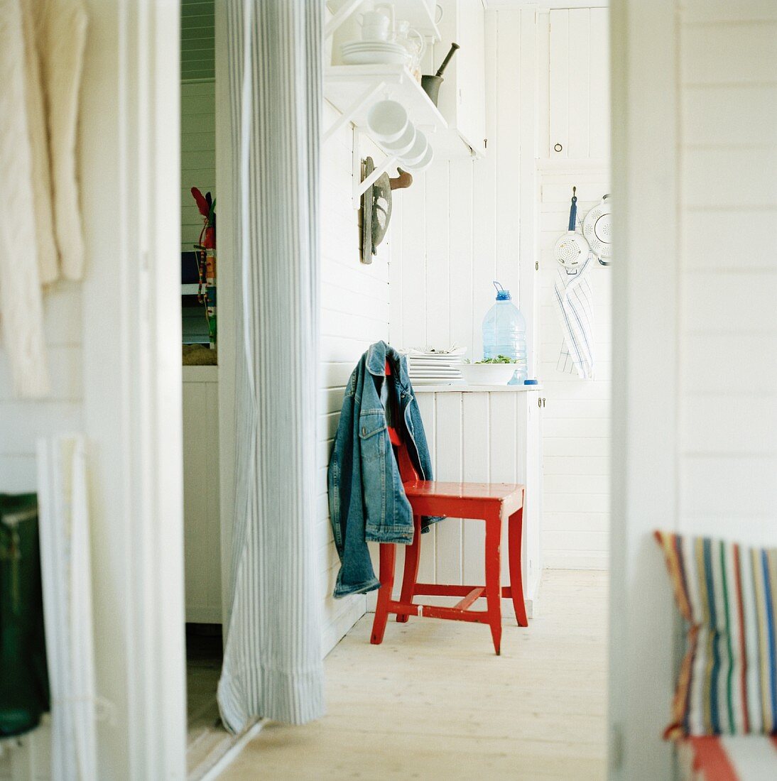 A red lacquered kitchen chair with a jacket hanging over the back next to a hallway with a white curtain