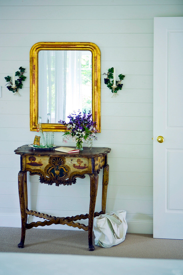 Antique, carved console table below gilt-framed mirror in rustic foyer