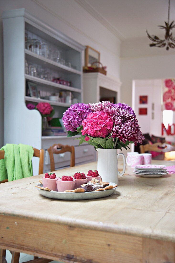 Pink and violet bouquet in ceramic jug and tray of desserts on rustic dining table