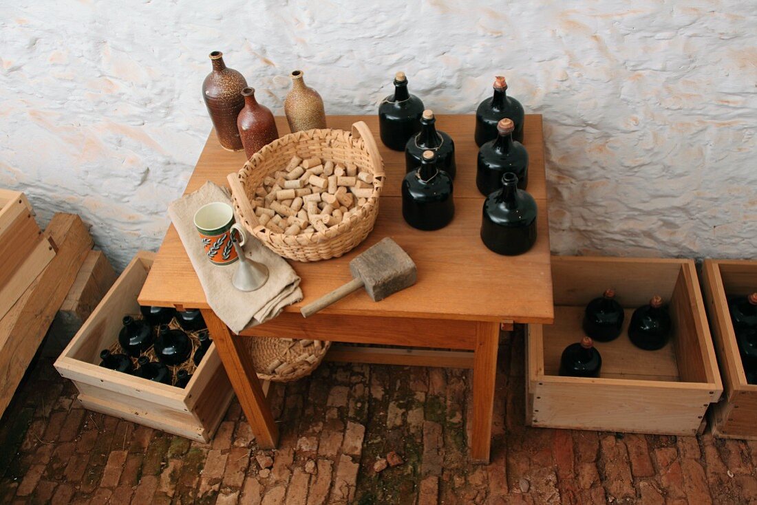 Various bottles made of clay and black ceramic next to a basket of corks on a wooden table